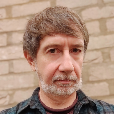 A white man with a grey beard wearing a check flannel shirt standing in front of a brick wall
